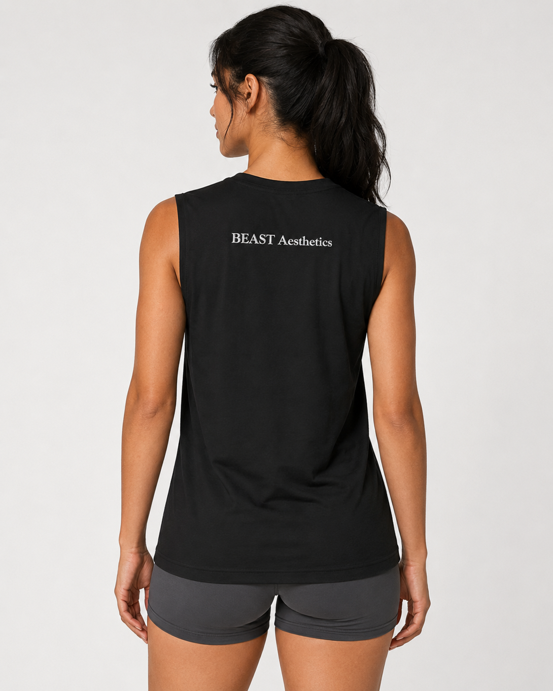 A woman shown from the back wearing a black sleeveless athletic tank top with 'BEAST Aesthetics' printed in white across the upper back, paired with dark gray athletic shorts, against a light gray studio background.