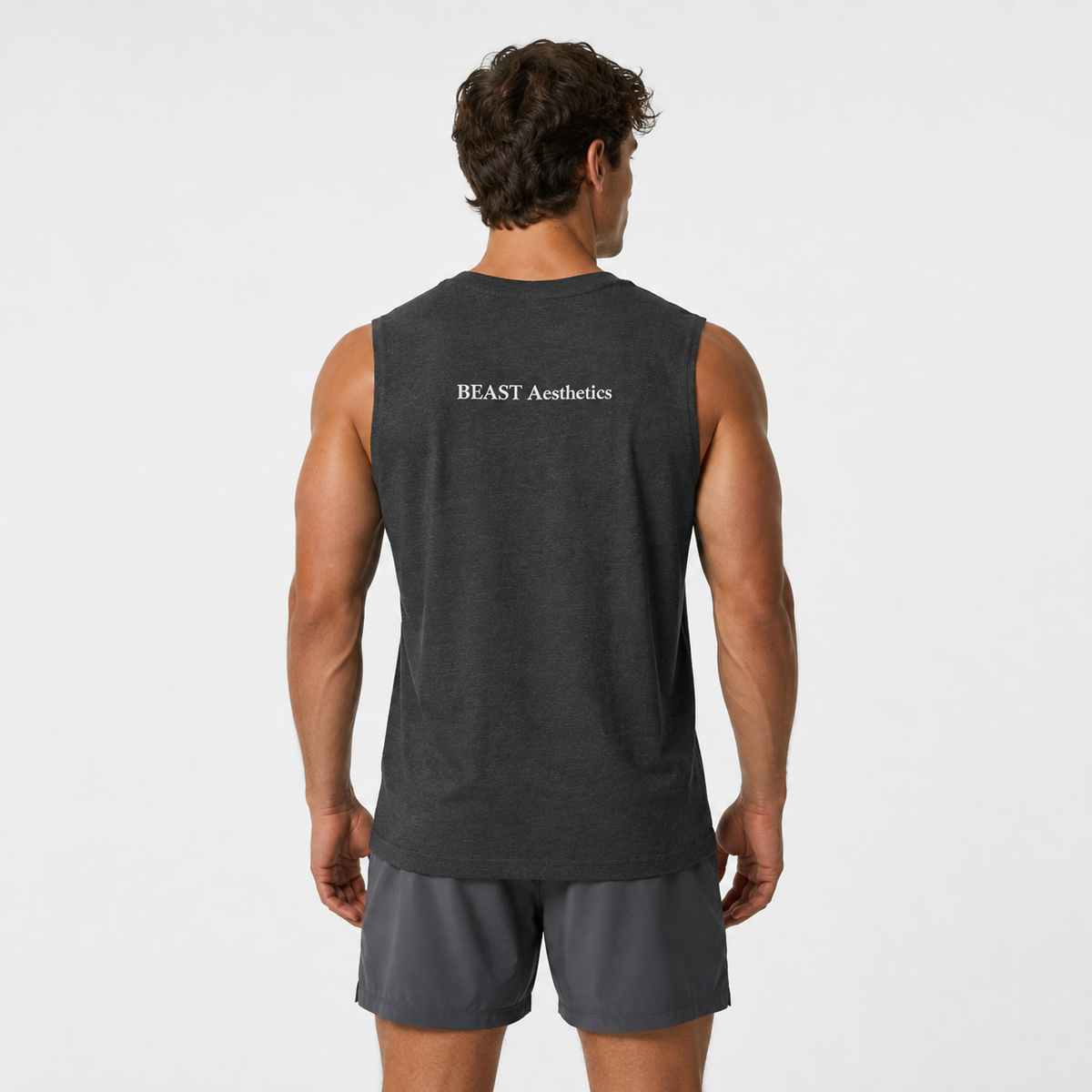 A man viewed from behind wearing a dark charcoal gray sleeveless athletic tank top with 'BEAST Aesthetics' printed in white on the upper back, paired with dark gray shorts, against a light gray studio background.