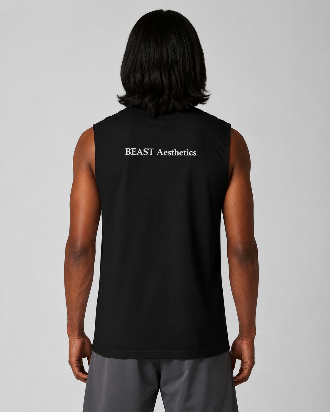 A man viewed from behind wearing a black sleeveless athletic tank top with 'BEAST Aesthetics' printed in white text on the back, paired with gray athletic shorts, against a solid gray studio background.