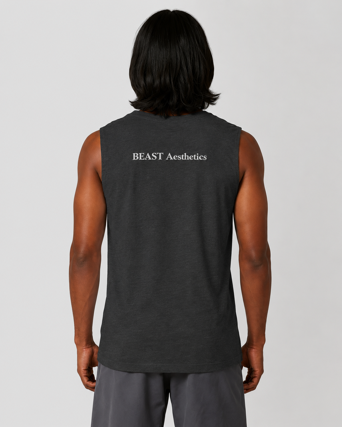 A man viewed from behind wearing a dark charcoal gray sleeveless muscle tank top with 'BEAST Aesthetics' printed in white text on the upper back, paired with gray sweatpants against a light gray studio background.
