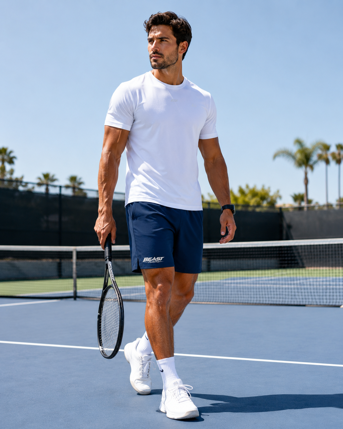 A man walking on a blue tennis court wearing a white t-shirt, navy blue athletic shorts with a white logo, white socks, and white sneakers, holding a tennis racket.