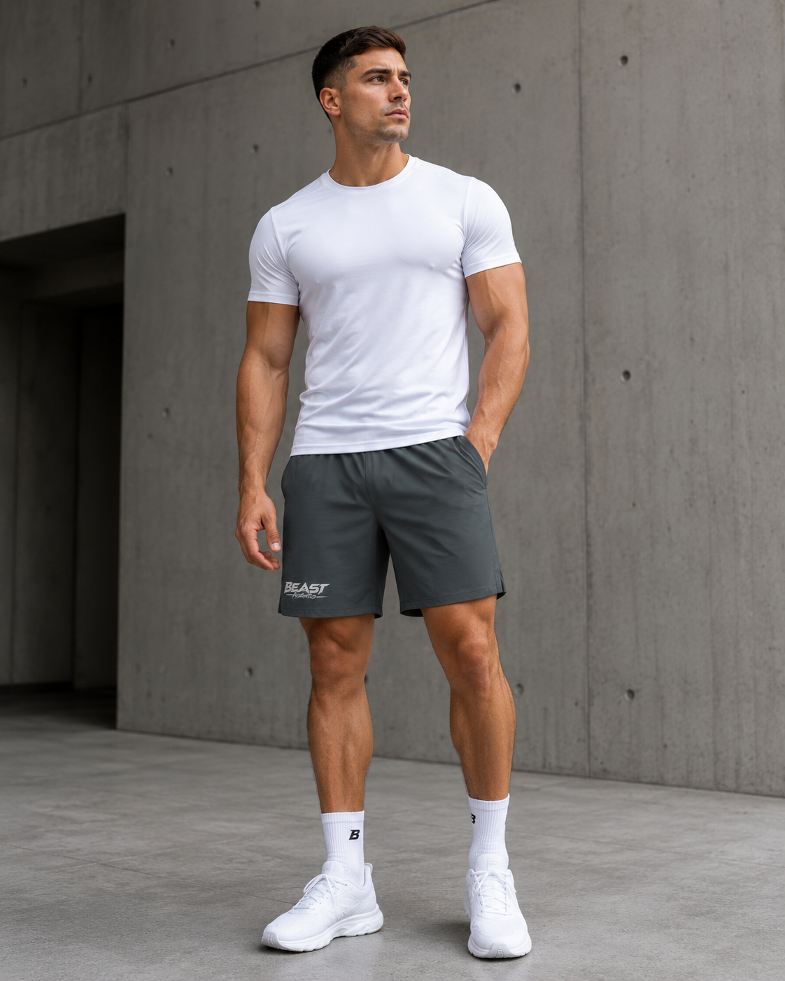 A man wearing a white t-shirt, dark gray athletic shorts with a white 'BLAZ' logo, white socks, and white sneakers, standing against a concrete wall.