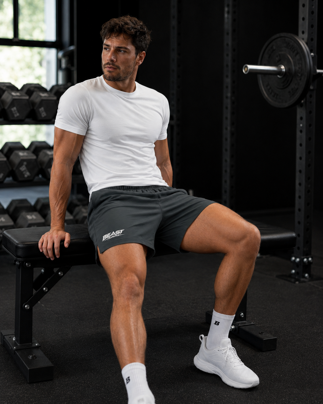 A man in a gym setting wearing a white t-shirt, dark gray athletic shorts with a white logo, white socks, and white sneakers, sitting on a weight bench.