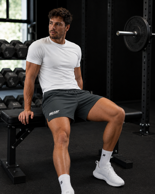 A man in a gym setting wearing a white t-shirt, dark gray athletic shorts with a white logo, white socks, and white sneakers, sitting on a weight bench.