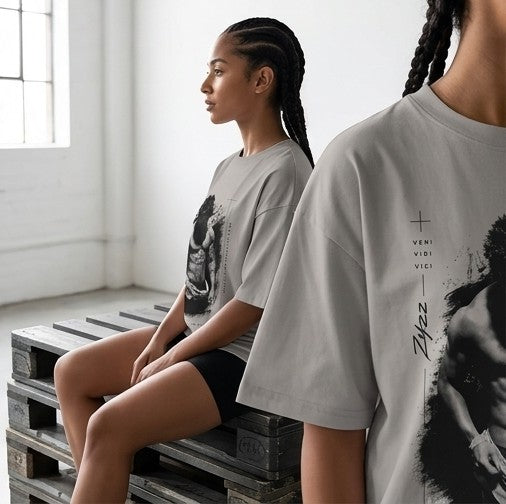A woman wearing a light gray oversized T-shirt with a black graphic print sits on wooden pallets in a bright room. Another person in the foreground wears the same shirt, showing the front print area.