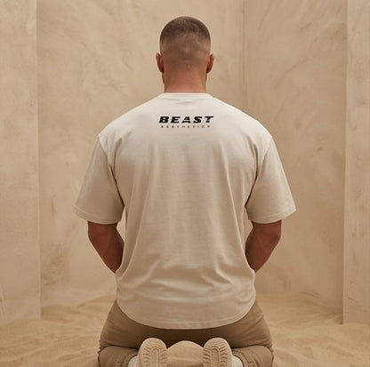 A man viewed from behind wearing a light beige t-shirt with 'BEAST AESTHETICS' printed in black on the back, kneeling in a minimalist beige room.