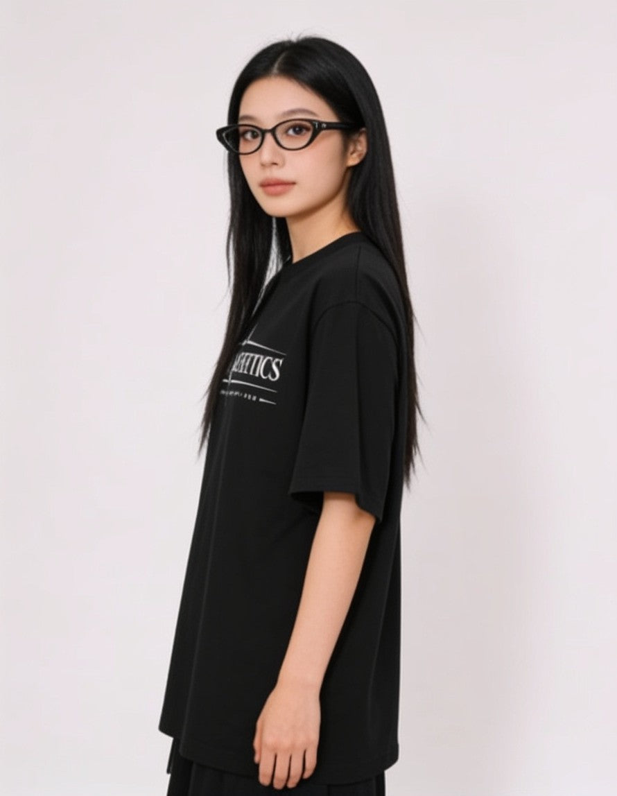 A woman with long black hair and black cat-eye glasses is shown in profile wearing an oversized black t-shirt with a white graphic print on the left chest, set against a plain light gray background.