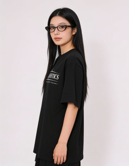 A woman with long black hair and black cat-eye glasses is shown in profile wearing an oversized black t-shirt with a white graphic print on the left chest, set against a plain light gray background.