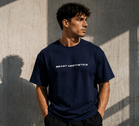 A man wearing a navy blue crewneck t-shirt with 'BEAST AESTHETICS' printed in white across the chest, standing against a textured concrete wall with dramatic shadows.