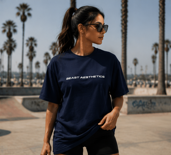 A woman wearing a navy blue t-shirt with 'BEAST AESTHETICS' printed in white across the chest, standing in an outdoor skatepark with palm trees in the background.