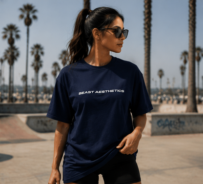 A woman wearing a navy blue t-shirt with 'BEAST AESTHETICS' printed in white across the chest, standing in an outdoor skatepark with palm trees in the background.