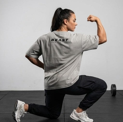 A woman in a gray t-shirt and black pants poses in a lunge position, flexing her arm. The back of the gray t-shirt features the text 'BEAST ATHLETICS' in black.