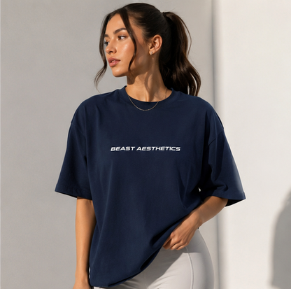 A woman wearing an oversized navy blue t-shirt with 'BEAST AESTHETICS' printed in white across the chest, paired with light gray leggings, set against a minimalist gray background.