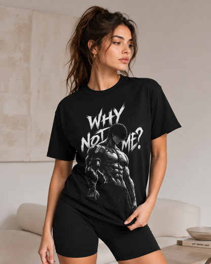 A woman modeling a black graphic t-shirt with the text 'WHY NOT ME?' and a muscular figure print, paired with black bike shorts in a modern indoor setting.