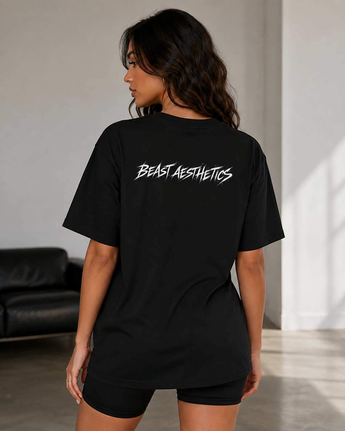 A woman wearing a black oversized t-shirt with 'BEAST AESTHETICS' printed in white across the back, shown from behind in a modern indoor setting.