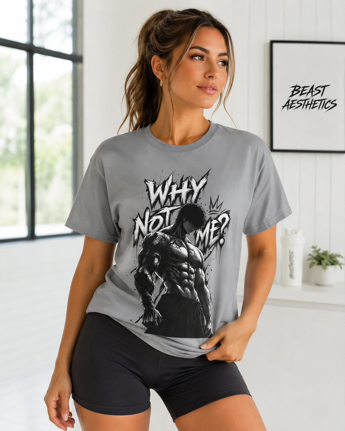 A woman modeling a gray graphic t-shirt with a black and white muscular man design and the text 'WHY NOT ME?' in a gym setting with a white paneled wall and a framed 'BEAST AESTHETICS' print.