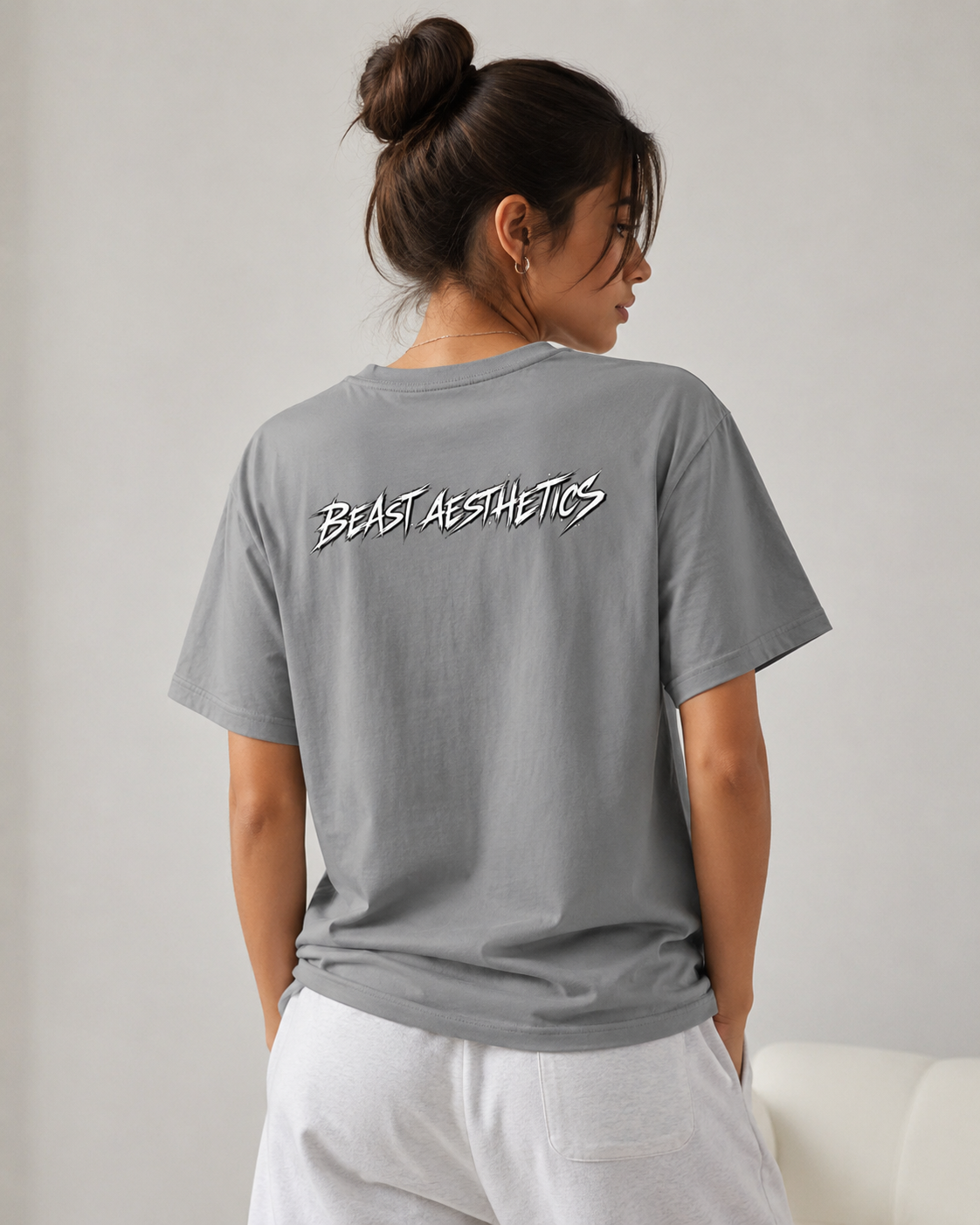 A woman shown from the back wearing a gray t-shirt with 'BEAST AESTHETICS' printed in white across the upper back, paired with white pants, set against a neutral indoor background.
