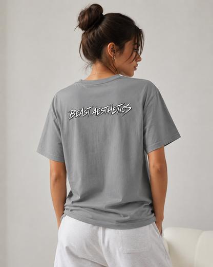 A woman shown from the back wearing a gray t-shirt with 'BEAST AESTHETICS' printed in white across the upper back, paired with white pants, set against a neutral indoor background.