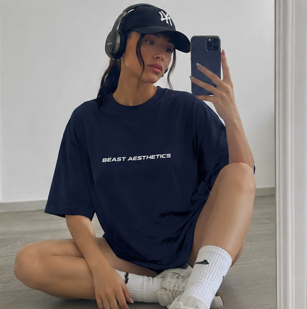A woman sitting on a floor in a mirror selfie wearing an oversized navy blue t-shirt with 'BEAST AESTHETICS' printed in white across the chest, a black baseball cap with a white NY logo, black headphones, white socks, and white sneakers.