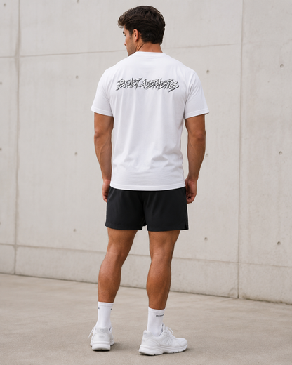 A man viewed from behind wearing a white t-shirt with a black graphic print on the back, black athletic shorts, white socks, and white sneakers, standing against a concrete wall.