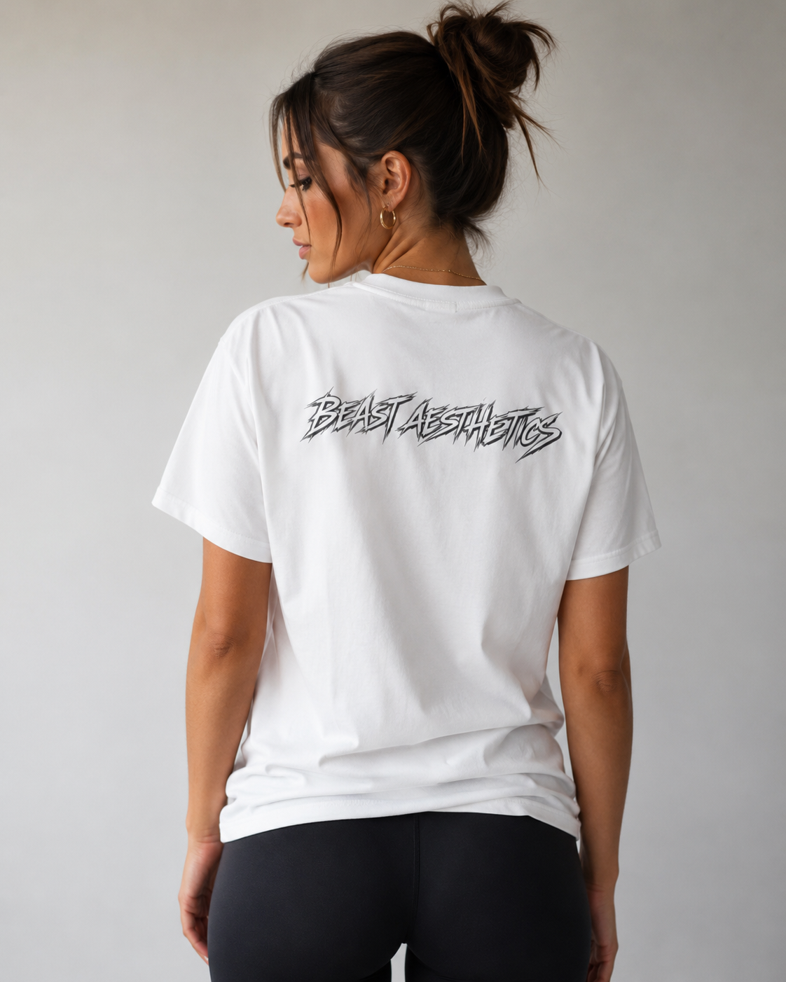 A woman shown from the back wearing a white short-sleeve T-shirt with a black 'BEAST AESTHETICS' graphic across the upper back, paired with black leggings, against a plain light gray background.