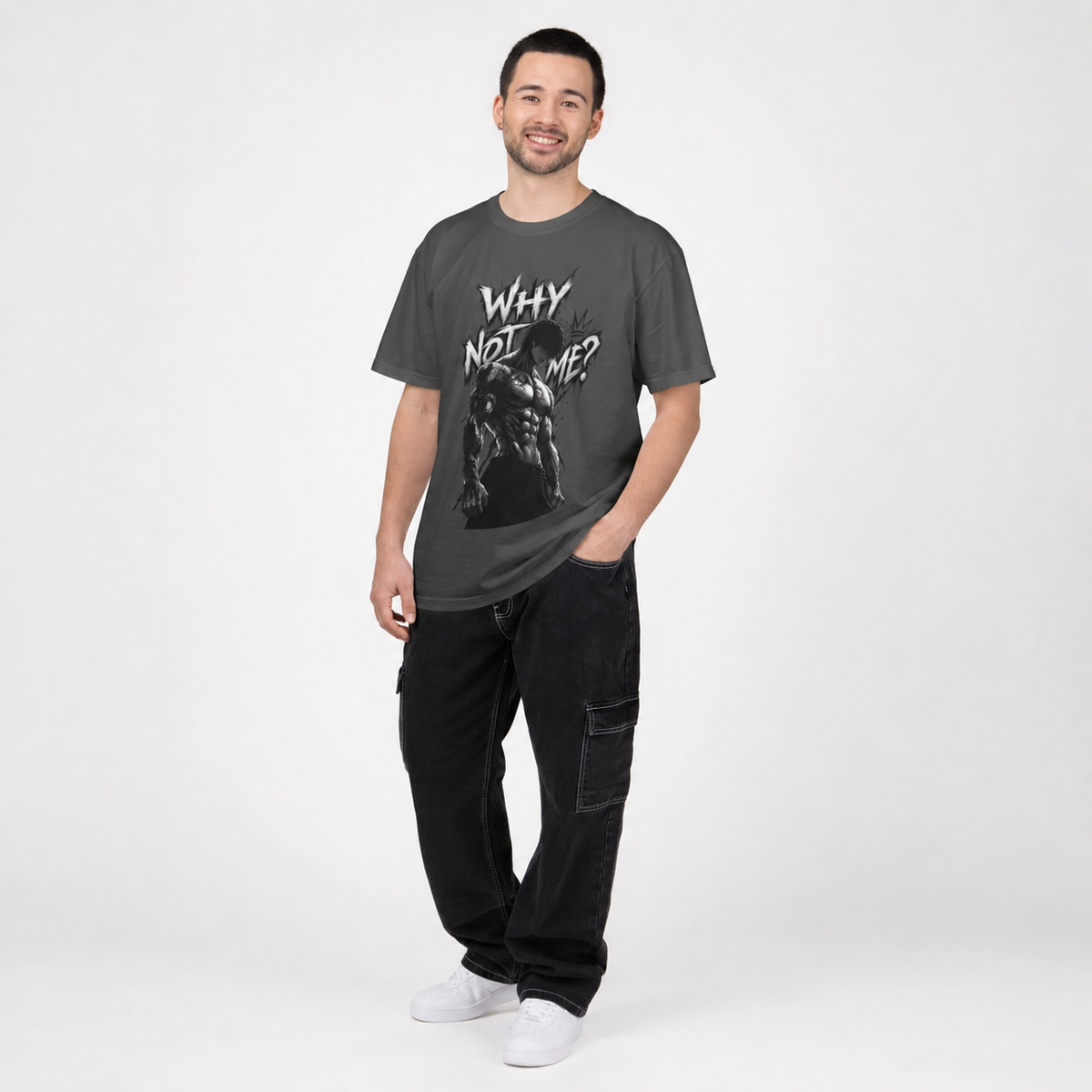 A man modeling a dark gray graphic t-shirt with the text 'WHY NOT ME?' and a black-and-white illustration, paired with black cargo pants and white sneakers, against a light gray studio background.
