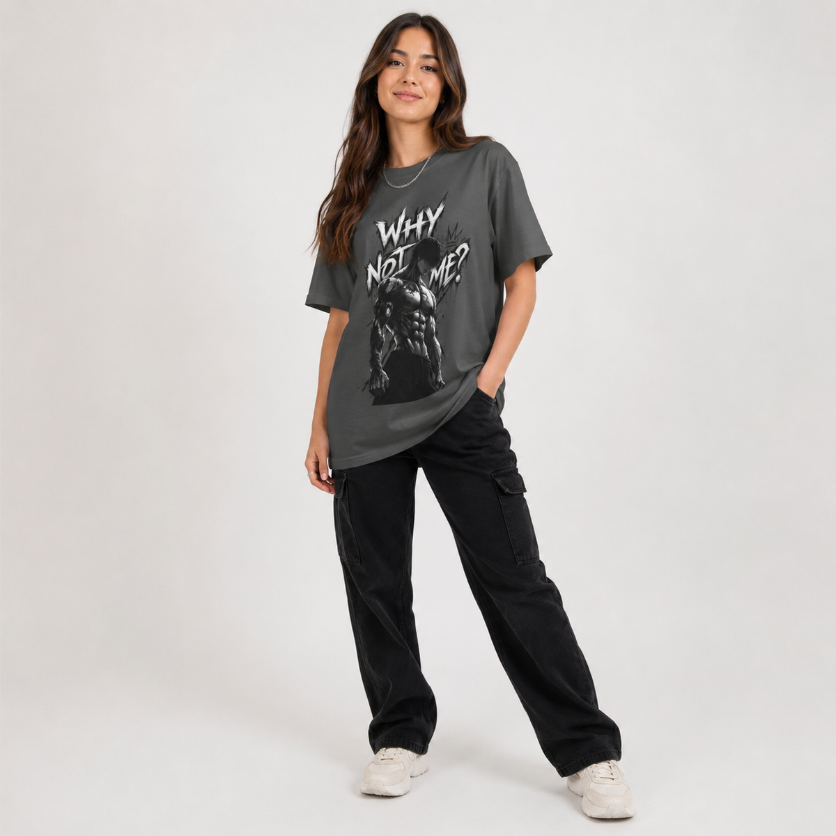 A woman modeling an oversized dark gray graphic t-shirt with the text 'WHY NOT ME?' and a black-and-white graphic print, paired with black cargo pants and white sneakers, against a light gray studio background.