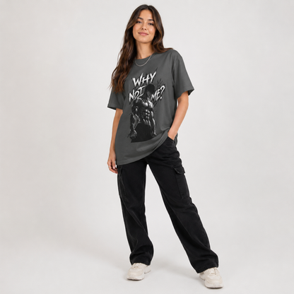 A woman modeling an oversized dark gray graphic t-shirt with the text 'WHY NOT ME?' and a black-and-white graphic print, paired with black cargo pants and white sneakers, against a light gray studio background.