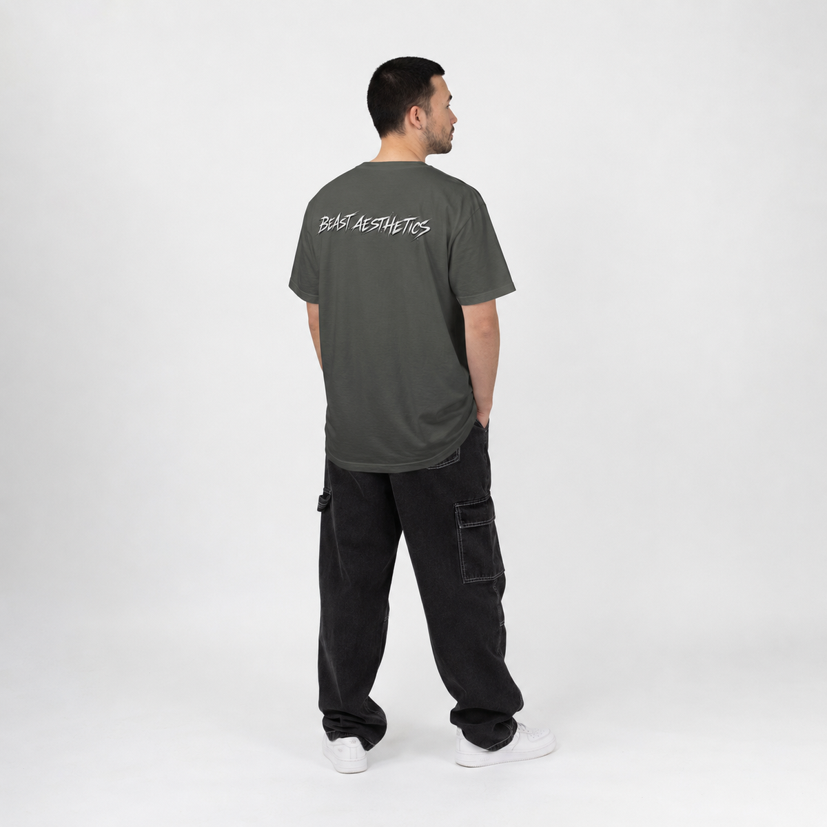 A man stands with his back to the camera wearing a dark gray t-shirt with 'BASE AESTHETICS' printed in white across the upper back, paired with black cargo pants and white sneakers, against a plain light gray studio background.