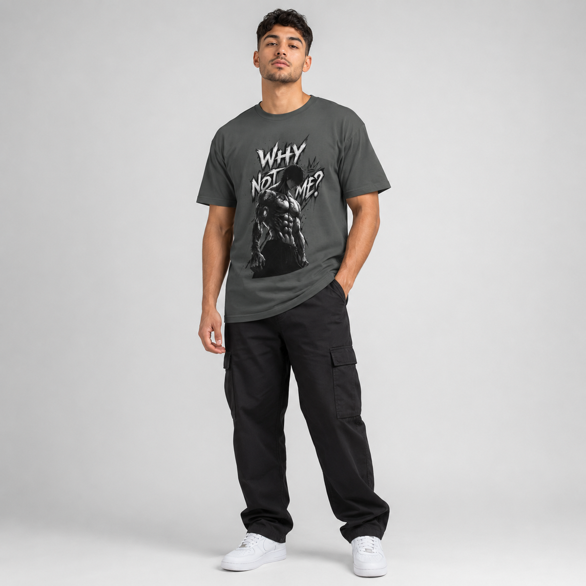 A man modeling a dark gray graphic t-shirt with the text 'WHY NOT ME?' and a muscular figure print, paired with black cargo pants and white sneakers, against a light gray studio background.