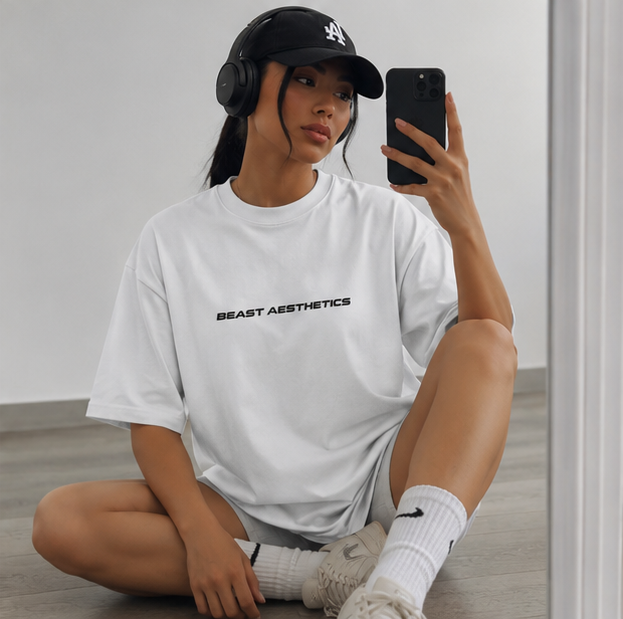 A woman sitting on a wooden floor takes a mirror selfie wearing an oversized white t-shirt with 'BEAST AESTHETICS' printed in black across the chest, a black baseball cap, black headphones, white socks, and white sneakers.