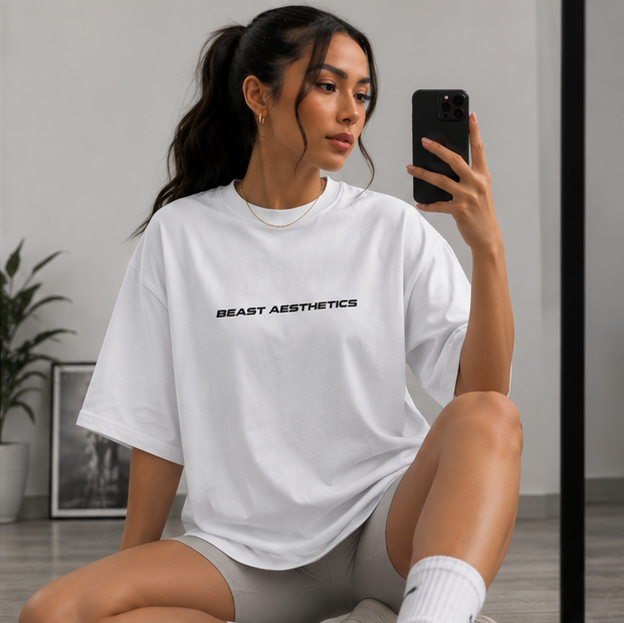 A woman takes a mirror selfie wearing a white oversized t-shirt with 'BEAST AESTHETICS' printed in black across the chest, paired with gray athletic shorts and white socks.