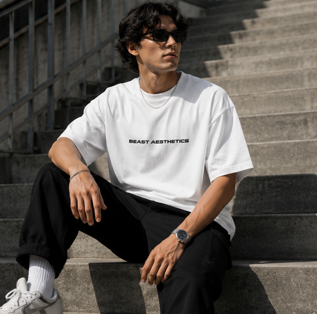 A man wearing a white short-sleeve t-shirt with 'BEAST AESTHETICS' printed in black across the chest, paired with black pants and white sneakers, sitting on concrete stairs.