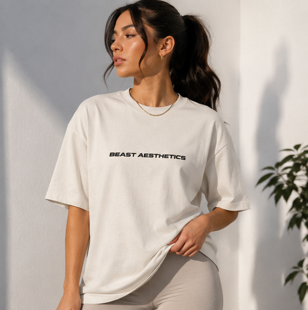 A woman wearing an oversized off-white t-shirt with 'BEAST AESTHETICS' printed in black across the chest, paired with light gray leggings, standing against a white wall with soft shadows.