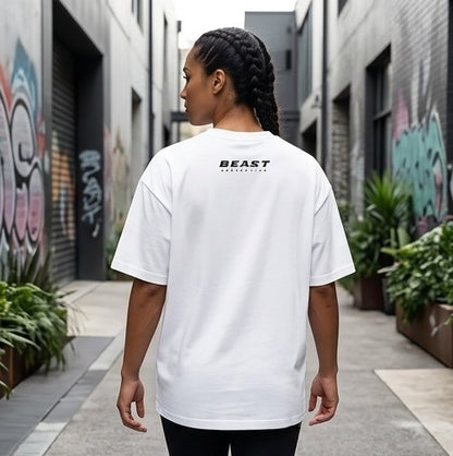 A person viewed from behind wearing an oversized white T-shirt with 'BEAST' printed in black across the upper back, standing in a narrow outdoor alley with graffiti walls and potted plants.