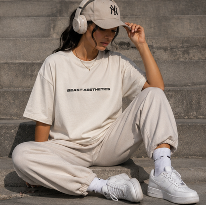 A woman sits on concrete steps wearing a beige NY cap, white over-ear headphones, a cream t-shirt with 'BEAST AESTHETICS' printed in black, matching cream sweatpants, white socks, and white sneakers.