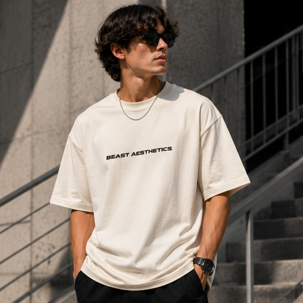 A man wearing a cream-colored t-shirt with 'BEAST AESTHETICS' printed in black across the chest, standing outdoors against a concrete wall and staircase.