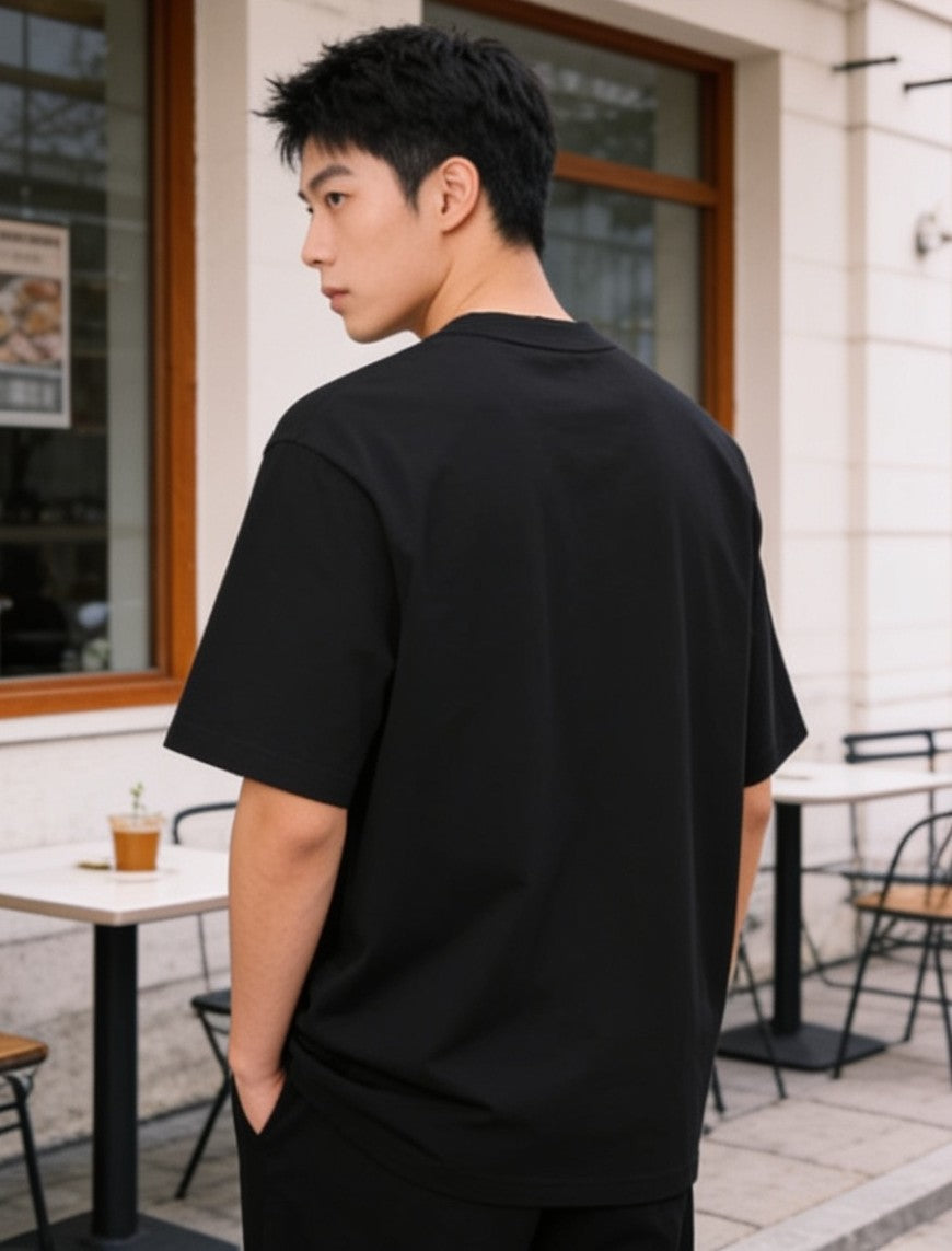A man wearing an oversized black short-sleeve t-shirt, photographed from behind in an outdoor cafe setting.