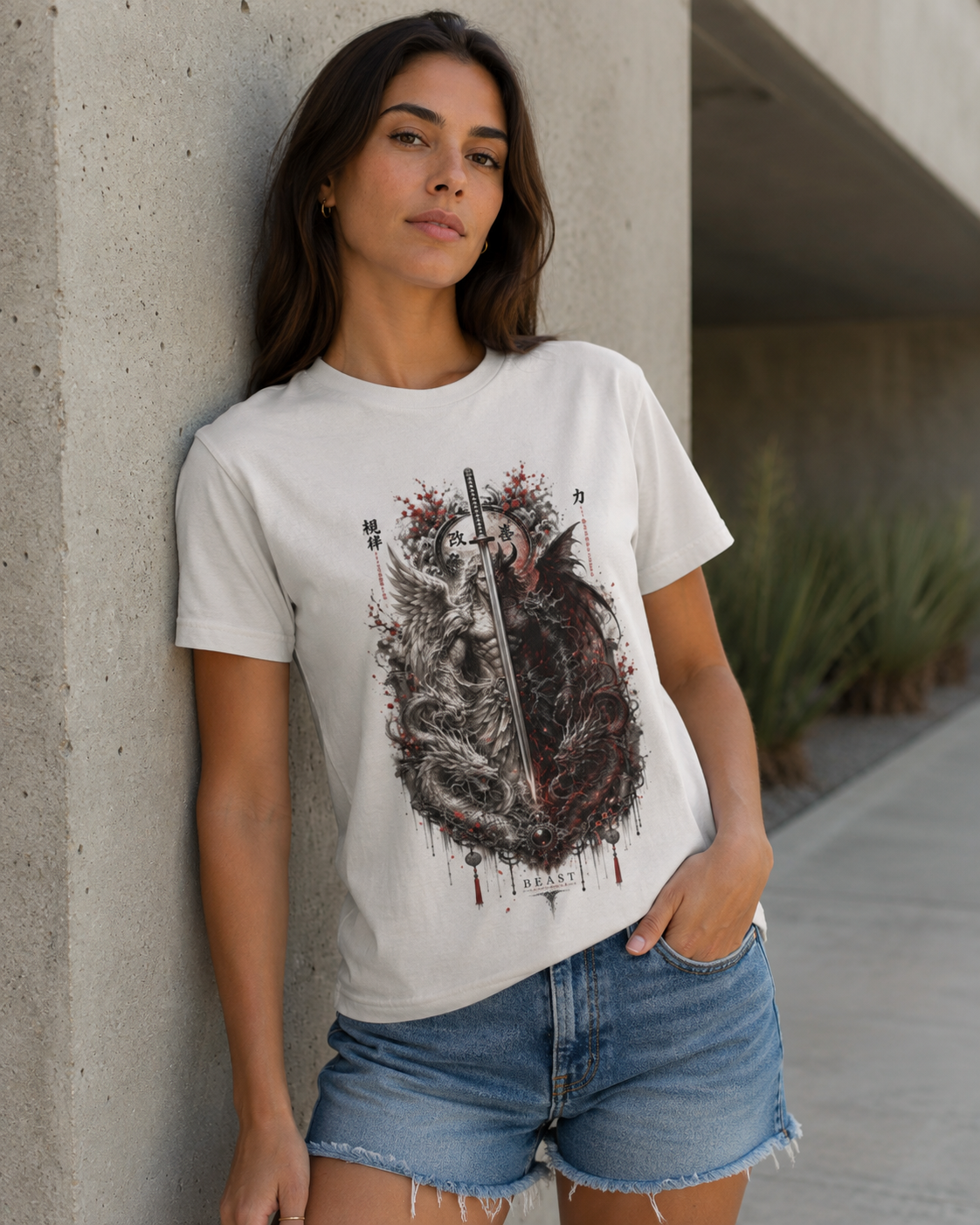 A woman wearing a white graphic t-shirt with a detailed gothic illustration of a heart, dagger, and playing cards, paired with denim shorts, leaning against a concrete pillar.