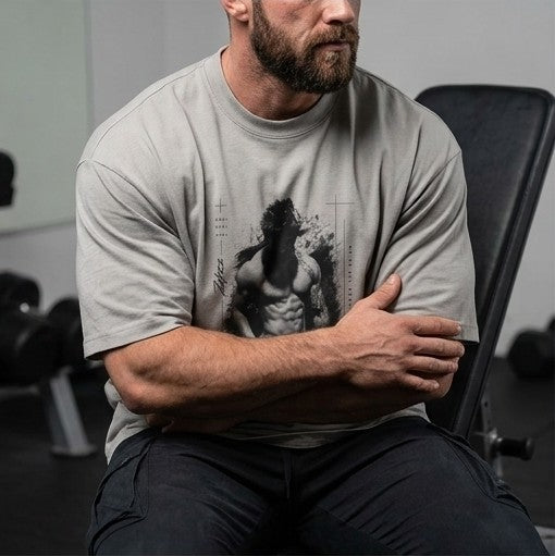 A man in a gym wearing a light gray t-shirt with a black graphic print of a muscular man, sitting on a weight bench.