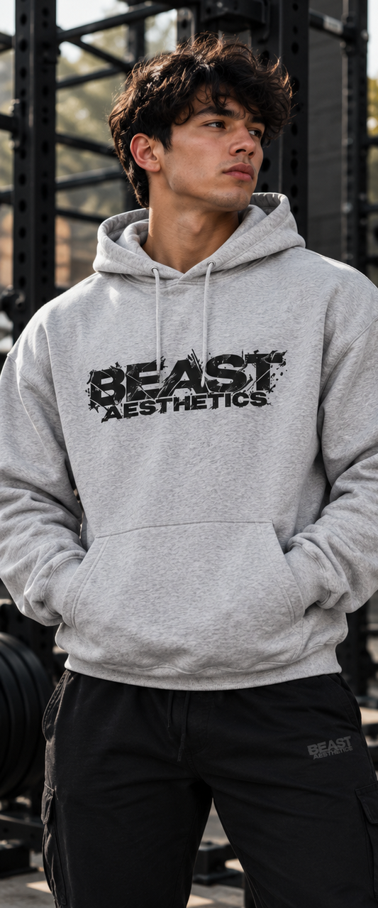 A man wearing a light gray heather hoodie with a black distressed 'BEAST AESTHETICS' logo on the chest, standing in a gym environment.