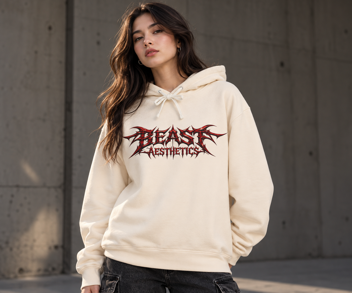 A woman wearing a cream-colored hoodie with a red and black 'BEAST AESTHETICS' graphic on the chest, standing against a concrete wall.