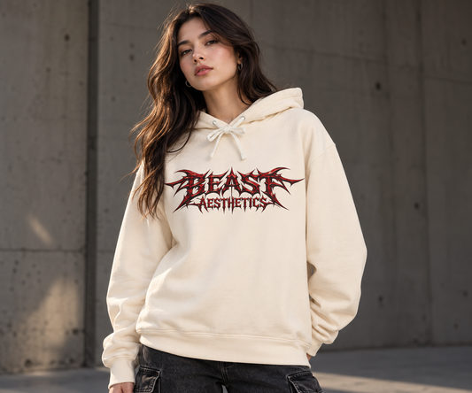 A woman wearing a cream-colored hoodie with a red and black 'BEAST AESTHETICS' graphic on the chest, standing against a concrete wall.