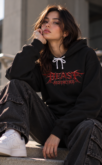 A woman sits on concrete steps wearing a black hoodie with a red and white 'BEAST AESTHETICS' graphic, paired with dark gray cargo jeans and white sneakers.