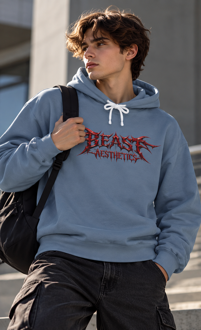 A young man wearing a dusty blue hoodie with a red and black 'BEAST AESTHETICS' graphic on the chest, black cargo pants, and a black duffel bag, standing outdoors against a gray concrete background.