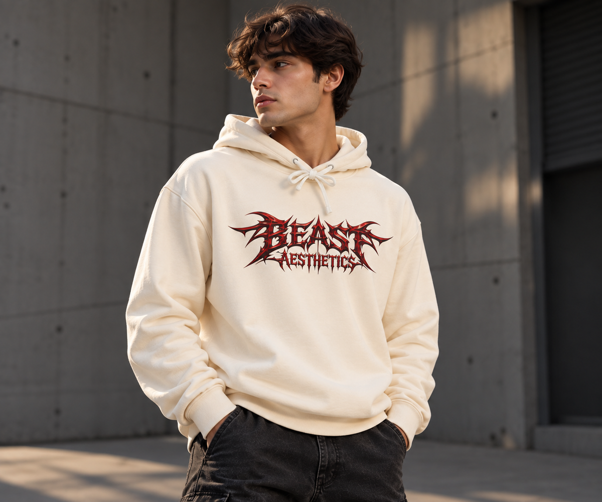 A man wearing a cream-colored hoodie with a red and black 'BLAST AESTHETICS' graphic on the chest, standing outdoors against a concrete wall.