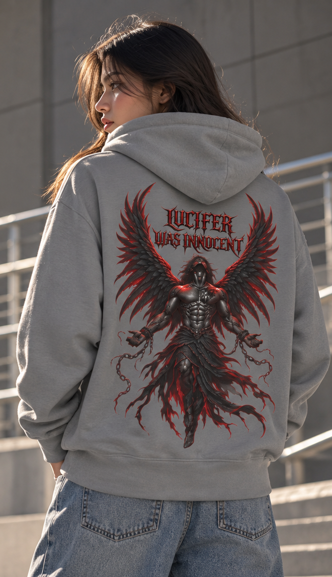 A woman viewed from behind wearing a light gray hoodie with a large red and black graphic of a winged demon and the text 'LUCIFER WAS INNOCENT' on the back, paired with blue jeans in an outdoor setting.