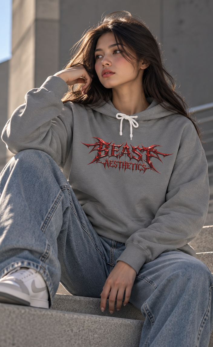 A woman sitting on concrete steps wearing a gray hoodie with a red and black 'BEAST AESTHETICS' graphic on the chest, paired with light blue jeans and white sneakers.