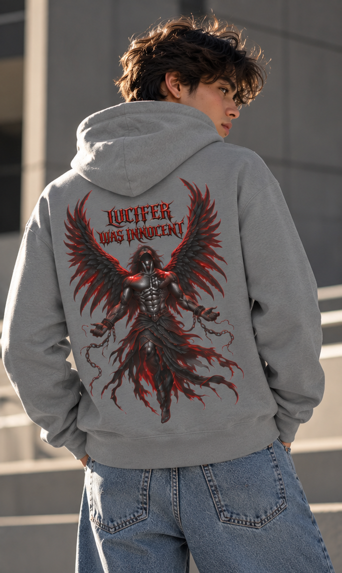 A person wearing a light gray hoodie from the back, featuring a large graphic print of a dark winged figure with red glowing accents and the text 'LUCIFER WAS INNOCENT'. The person is also wearing light blue jeans, set against a modern concrete building background.
