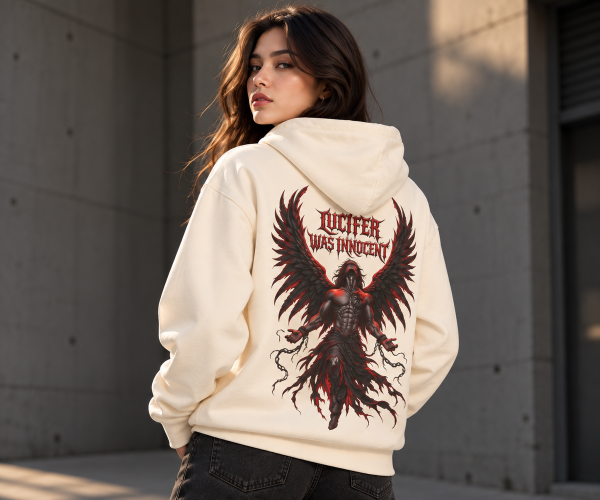 A woman wearing a cream-colored hoodie with a large back graphic of a red and black winged figure and the text 'LUCIFER WAS INNOCENT', photographed outdoors against a concrete wall.
